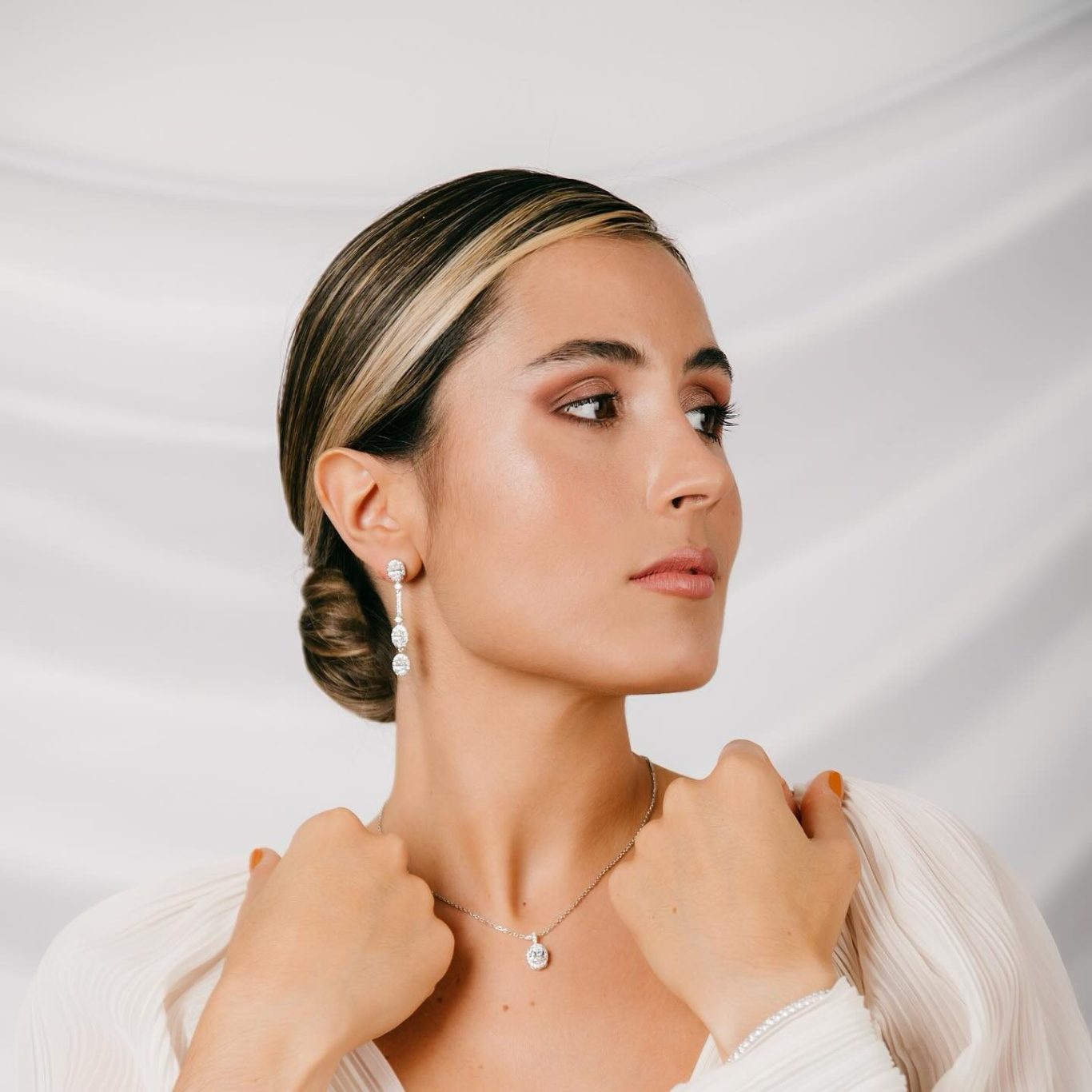 Complementos para novias: pendientes, anillos, colgantes Mujer con peinado elegante, usando pendientes y collar, con fondo de tela clara.